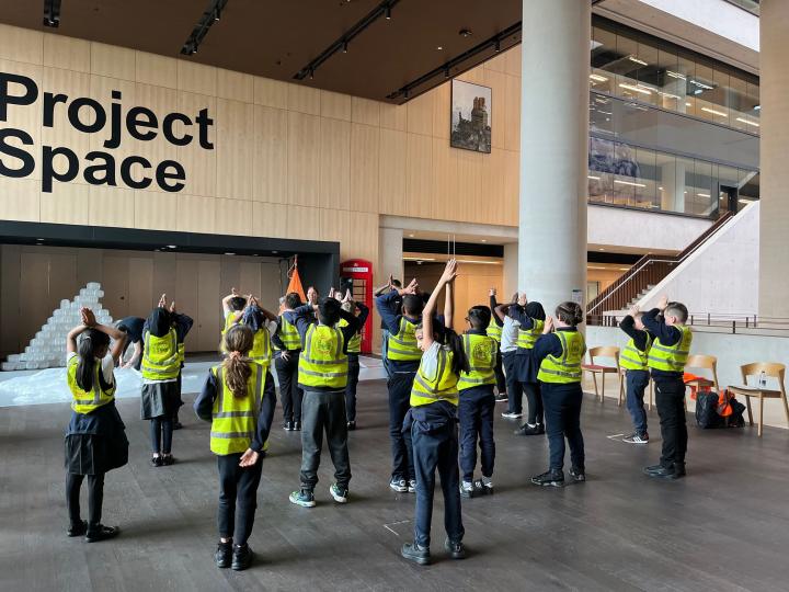 A group of school pupils in high vis jackets with their backs towards the camera, following arms movements as instructed by the artist facing them and the camera.